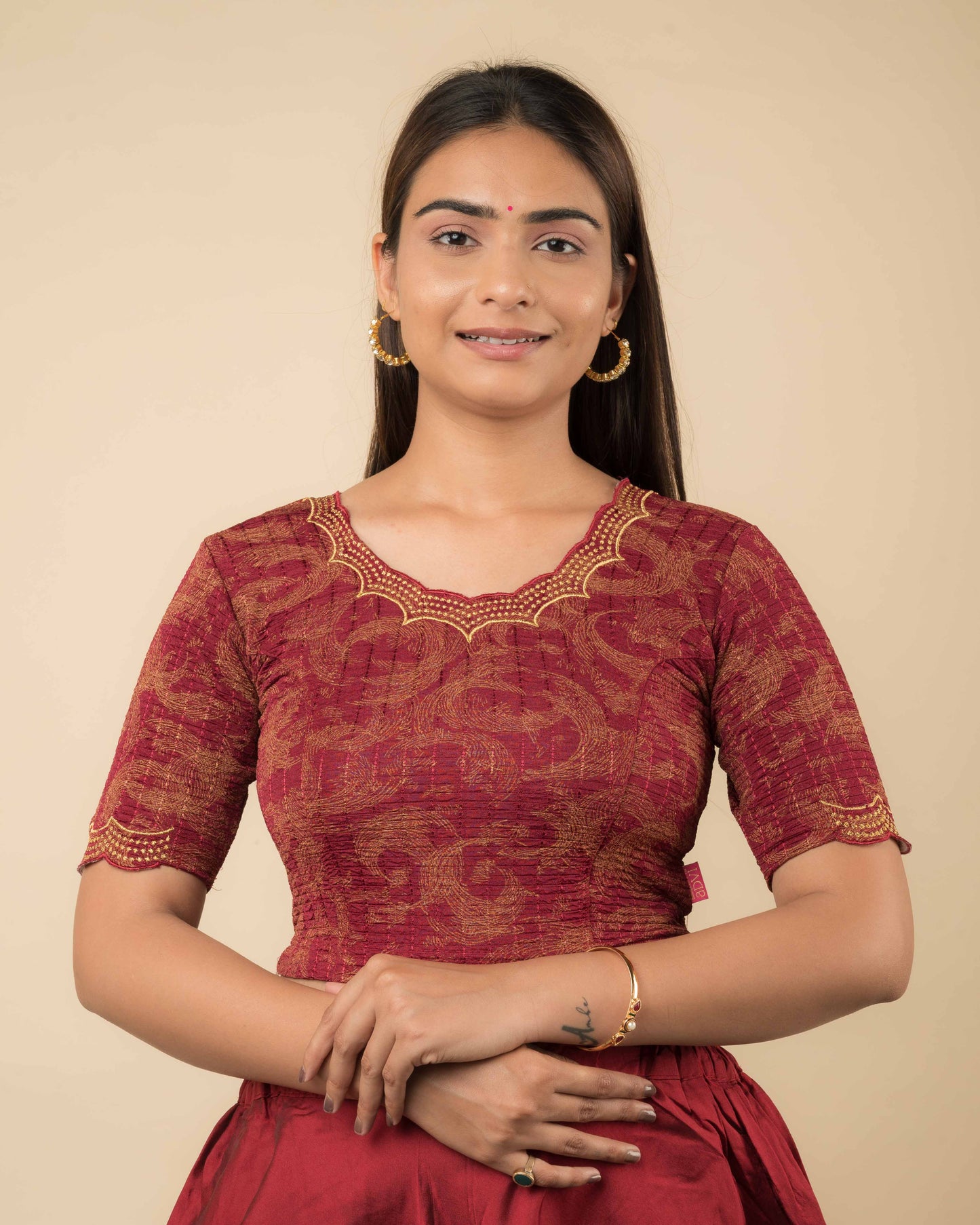 Model wearing a maroon-colored stretchable readymade blouse with zari embroidery, elbow sleeves, and scalloped round neckline.