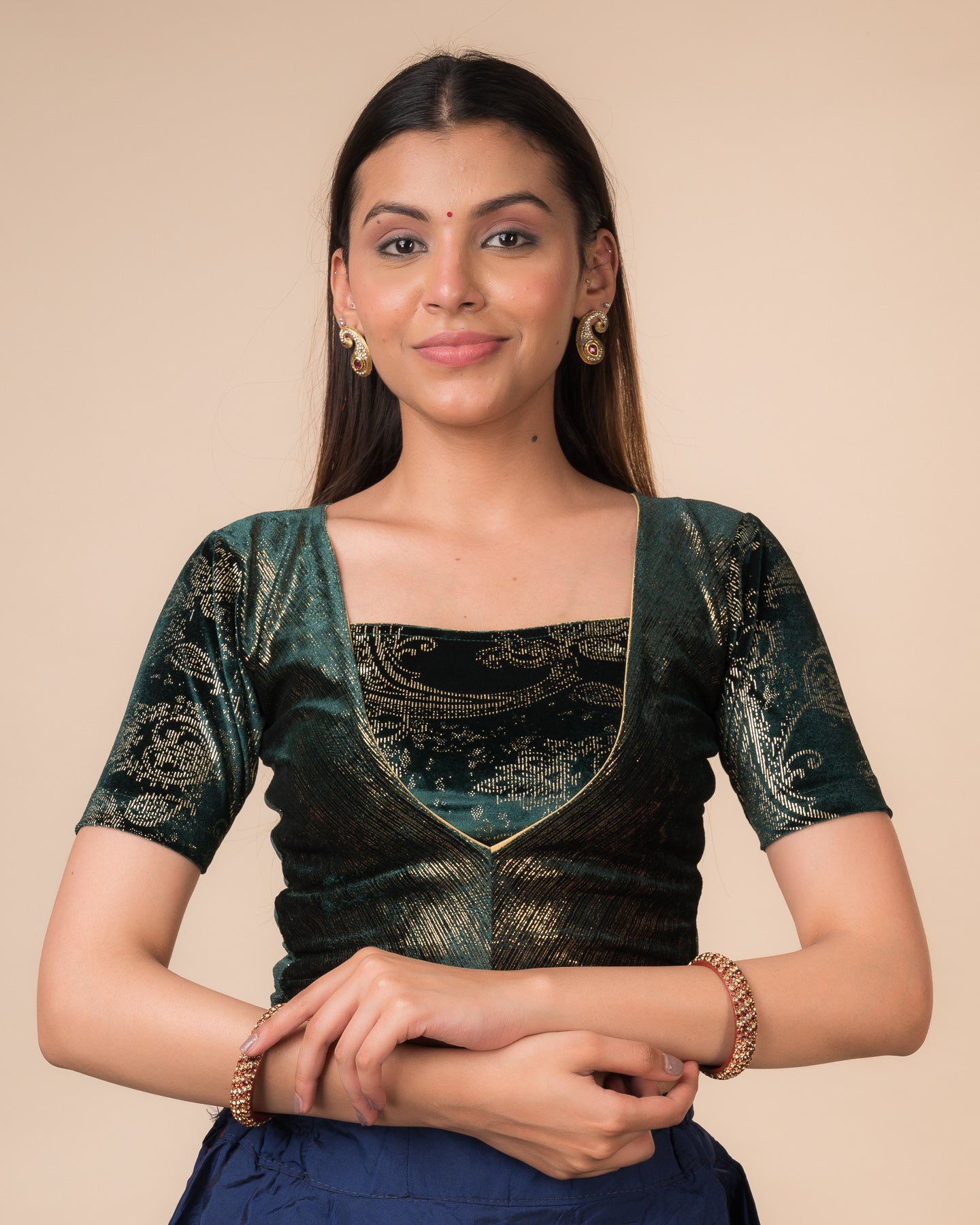Model wearing a bottle green-colored velvet embellished stretchable choli-style readymade blouse with elbow sleeves and round neckline.